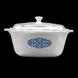 May include: A white rectangular casserole dish with a clear glass lid and integrated side handles. The front of the dish features a centred blue decorative filigree pattern. The vessel has rounded corners and a smooth, opaque finish throughout.