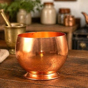 May include: A polished copper bowl with a rounded body and a flared circular base. The metal surface features a warm, reflective metallic finish with subtle texture and a wide, open top, showcasing a consistent reddish-orange hue throughout.