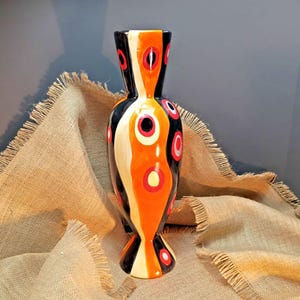 May include: A tall ceramic vase featuring a glossy finish with vertical wavy bands of black, orange, and cream. The surface is decorated with a repeating pattern of circular motifs in red, white, and black, arranged in a vertical, abstract design.