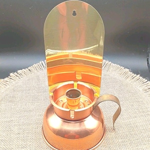 May include: A vintage copper candle holder with a handle and a curved, reflective back. The candle holder has a round base and a cylindrical cup for the candle. The back is a curved, metallic shield. The candle holder is on a textured, neutral-colored surface.