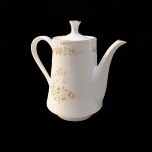 May include: A white porcelain teapot featuring a delicate floral pattern with pink and white roses, green leaves, and a small blue bird. The teapot has a curved handle, a tapered spout, and a matching lid with a small knob handle on top.