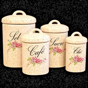 May include: A set of four cylindrical ceramic canisters with lids, featuring a speckled cream finish, arched handles, and floral accents. Each canister is labeled in black script with the words Sel, Café, Sucre, and Thé, respectively.