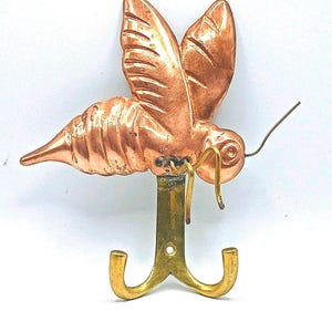 May include: A copper-colored metal wall hook shaped like a stylized insect with wings. The hook has a brass-colored base with two curved hooks. The insect has detailed wings and a segmented body, with a wire-like antenna.
