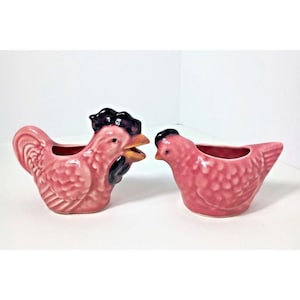 May include: Two pink ceramic chicken-shaped containers. One is a rooster with a black comb and beak, the other is a hen. The containers are likely for holding small items or used as decorative pieces.