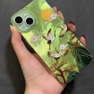 Peut inclure: Coque de téléphone verte avec un motif de grue. L'étui présente un fond vert texturé avec un soleil doré, des nuages blancs et des grues en vol. Le motif comprend un arbre et une chaîne de montagnes.