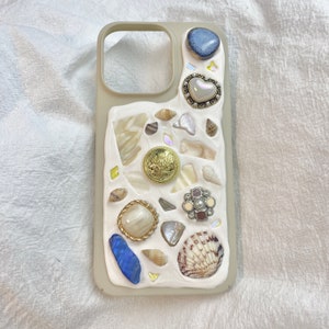 May include: A beige iPhone case decorated with seashells, pearls, and gold accents. The case has a textured surface and a variety of different sized and shaped shells.