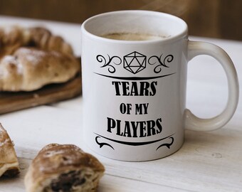 Op de afbeelding: Witte keramische mok met de tekst "TEARS OF MY PLAYERS" in zwart, met een D20-afbeelding. De mok is gevuld met een warme drank, met stoom die opstijgt. Croissants liggen op een houten plank.