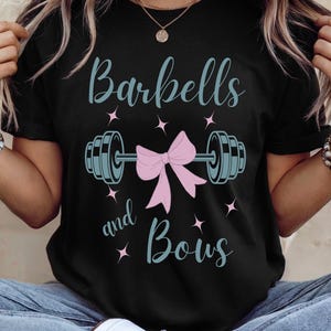 May include: Black t-shirt with the text "Barbells and Bows" in light blue script. A barbell graphic is centered, with a pink bow in the middle. Pink star accents surround the text and graphic.