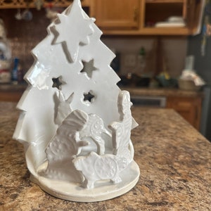 Peut inclure: Ornement de Noël en céramique blanche avec une scène de la nativité à l'intérieur. L'arbre a des étoiles découpées et la scène de la nativité comprend Marie, Joseph et l'enfant Jésus.