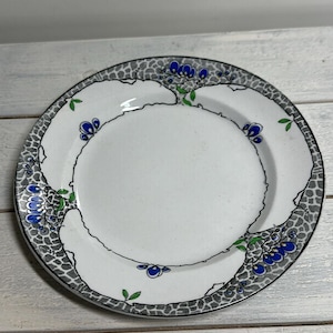 Puede incluir: Plato de cerámica blanca con un diseño floral gris, negro, azul y verde. El plato tiene un borde festoneado con un patrón gris. El diseño floral incluye racimos de flores azules y hojas verdes.