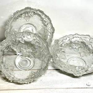 May include: Three clear glass bowls with floral and leaf designs. The bowls have a scalloped edge and a circular base. The glass has a frosted appearance. The bowls are arranged on a white wooden surface.