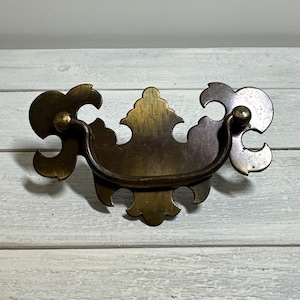 May include: Antique brass drawer pull with a decorative, ornate design. The pull features a curved handle and intricate detailing on the backplate. The hardware is a warm, aged brass color, adding a vintage touch to furniture.