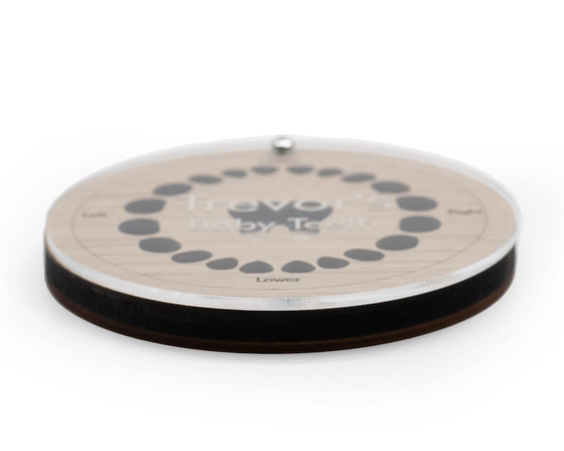 May include: A wooden baby teeth tracker with a clear acrylic cover. The tracker has a circle of 20 small black circles to mark the baby teeth. The text "Travo Baby Teeth" is printed on the tracker. The text "Lower" is printed below the circle of black circles.