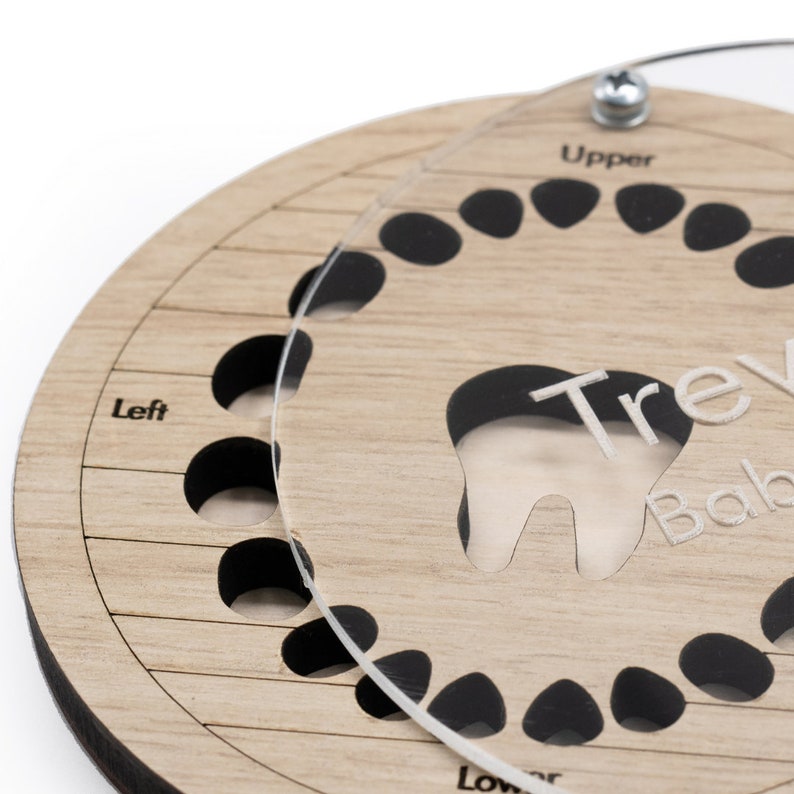 May include: A wooden baby tooth keepsake box with a clear acrylic lid. The box has a circular shape and features a tooth-shaped cutout with the name "Trevor" engraved on it. The box has slots for storing baby teeth, labeled "Upper", "Lower", "Left", and "Right".