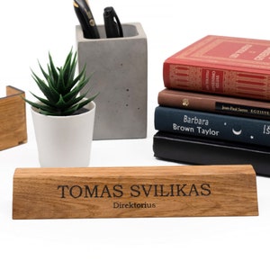 Op de afbeelding: Een houten bureau-naamplaatje met de tekst "TOMAS SVILIKAS Direktorius" gegraveerd in zwarte letters. Het naamplaatje ligt op een licht houten oppervlak.