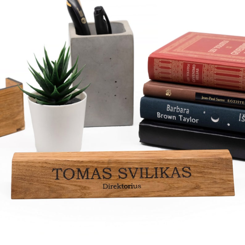 Op de afbeelding: Een houten bureau-naamplaatje met de tekst "TOMAS SVILIKAS Direktorius" gegraveerd in zwarte letters. Het naamplaatje ligt op een licht houten oppervlak.