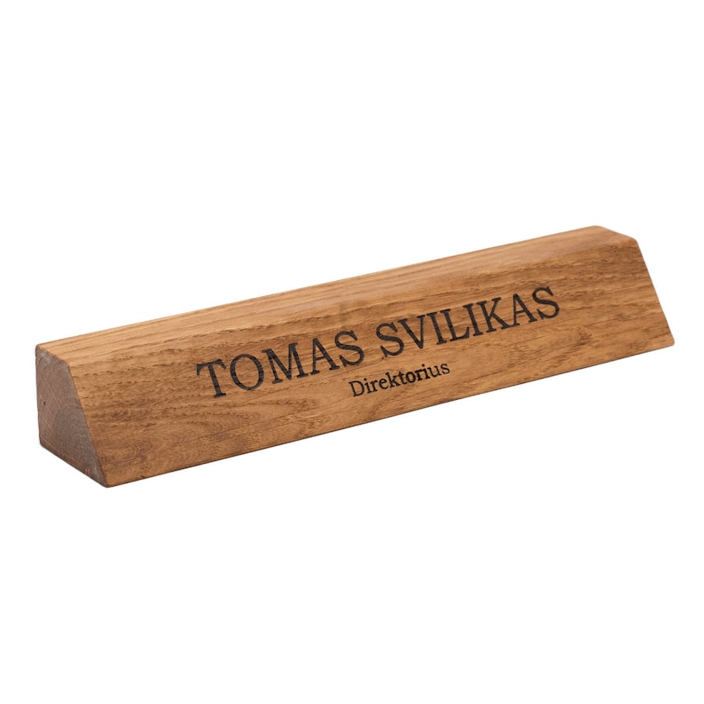 Op de afbeelding: Een houten bureau naamplaatje met de tekst "TOMAS SVILIKAS Direktorious" gegraveerd in zwart.