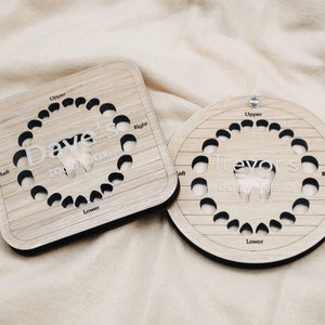 May include: Two wooden tooth boxes with a circular and square shape. The boxes are made of light brown wood with a wood grain pattern. The boxes have a cutout of a tooth in the center and are labeled with the names "Dave's Tooth Box" and "Trevor's Baby Teeth".