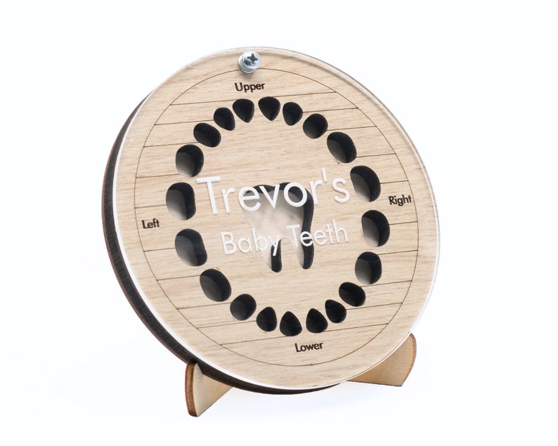 May include: A wooden baby teeth chart with a cutout of a tooth in the center. The chart is labeled "Trevor's Baby Teeth" and has spaces for 20 teeth. The chart is on a small wooden stand.