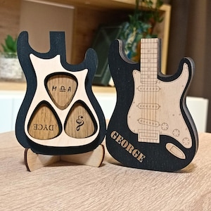 Könnte beinhalten: Zwei Gitarrenförmige Halter aus Holz. Ein Halter hat drei Fächer für Gitarrenplektren, beschriftet mit "DYCE", "MQA" und einer Musiknote. Der andere Halter hat einen schwarzen Hintergrund mit dem Wort "GEORGE" in weißen Buchstaben.