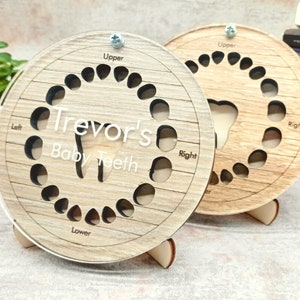 Puede incluir: Un gráfico de dientes de leche de madera con el nombre "Trevor's Baby Teeth" grabado en él. El gráfico tiene ranuras para los 20 dientes de leche y está etiquetado como "Upper", "Lower", "Left" y "Right".