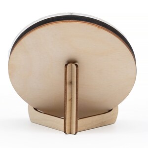 May include: A round wooden plaque with a stand. The plaque is light brown and has a dark brown edge. The stand is made of the same wood and has a triangular shape.