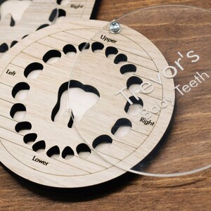 May include: A wooden baby teeth chart with a clear plastic cover. The chart has slots for each tooth and is labeled with "Upper", "Lower", "Left", and "Right". The cover has the name "Trevor's Baby Teeth" engraved on it.