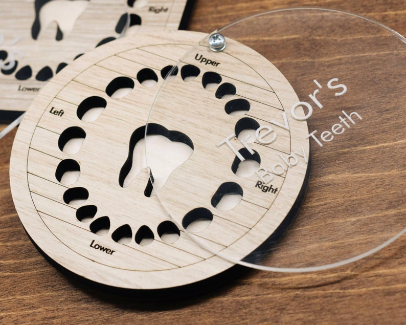 May include: A wooden baby teeth chart with a clear plastic cover. The chart has slots for each tooth and is labeled with "Upper", "Lower", "Left", and "Right". The cover has the name "Trevor's Baby Teeth" engraved on it.