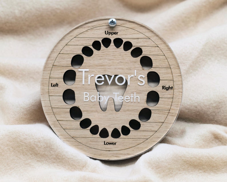 May include: A wooden baby teeth keepsake box with a circular design. The box has slots for all 20 baby teeth and is labeled "Trevor's Baby Teeth" with sections for "Upper", "Lower", "Left", and "Right".