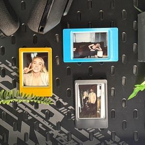 May include: A black pegboard wall organizer featuring three colorful magnetic photo frames in yellow, blue, and gray. The frames hold small rectangular photographs. The setup includes two gray wall-mounted planters with green plants and black headphones.