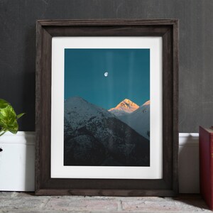 Puede incluir: Una fotograf&iacute;a enmarcada de una cordillera con luna llena en el cielo. Las monta&ntilde;as est&aacute;n cubiertas de nieve y el cielo es de un azul profundo.