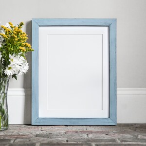 Puede incluir: Un marco de imagen azul claro con un paspart&uacute; blanco y un fondo blanco. El marco est&aacute; sentado en un suelo de ladrillo junto a un jarr&oacute;n de flores blancas y amarillas.