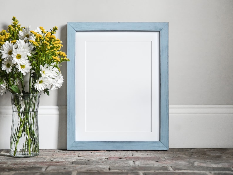 Puede incluir: Un marco de imagen azul claro con un paspart&uacute; blanco y un fondo blanco. El marco est&aacute; sentado en un suelo de ladrillo junto a un jarr&oacute;n de flores blancas y amarillas.