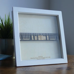 May include: A white framed print of a paper model of a building with the text "Mulgrave, Waterloo Road, Wolverhampton WV1 4GB, Capacity: 32,000" underneath.