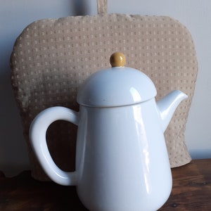 May include: White ceramic teapot with a yellow knob on the lid. The teapot is sitting on a wooden surface in front of a beige fabric tea cozy.