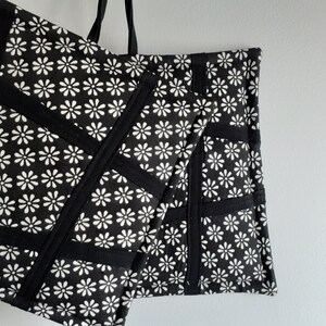 May include: Black and white tote bag with a daisy print and black fabric accents. The bag has a large capacity and is made of durable fabric.