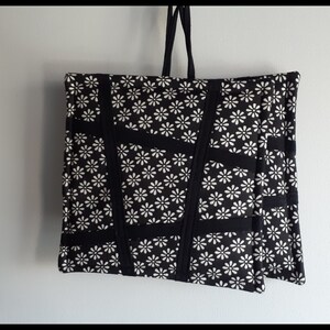 May include: Black tote bag with white daisy print and black straps. The bag is made of fabric and has a quilted design.