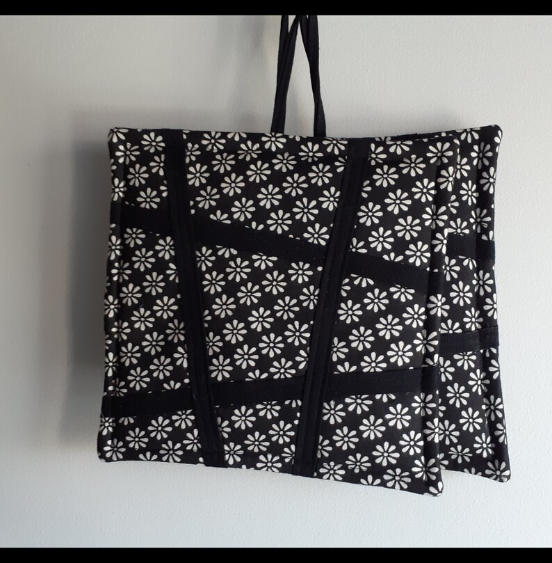 May include: Black tote bag with white daisy print and black straps. The bag is made of fabric and has a quilted design.