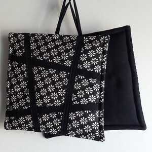 May include: Two black pot holders, one with a white floral pattern and the other plain. The pot holders are hanging from a white wall.