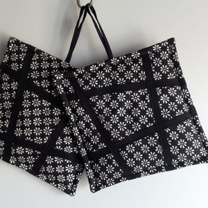 May include: Two black and white pot holders with a daisy print and black trim. The pot holders are hanging from a metal hook.