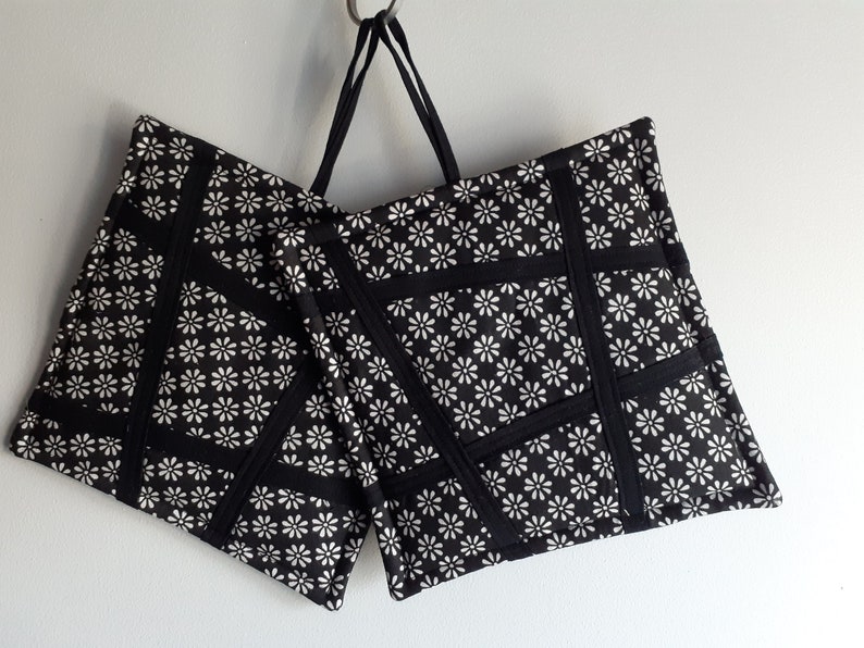 May include: Two black and white pot holders with a daisy print and black trim. The pot holders are hanging from a metal hook.