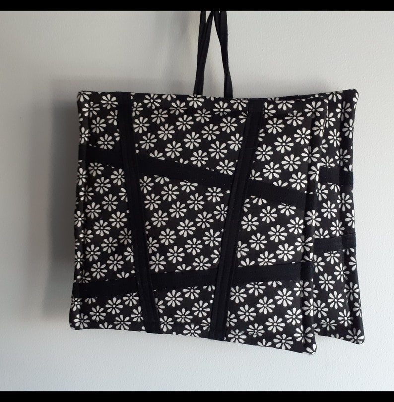 May include: Black tote bag with white daisy print and black straps. The bag is made of fabric and has a quilted design.