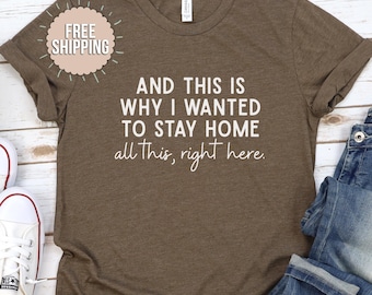 May include: A brown t-shirt with the text "AND THIS IS WHY I WANTED TO STAY HOME all this, right here." in white. The shirt has rolled-up sleeves and is paired with blue jeans and white sneakers. A "FREE SHIPPING" sticker is visible.