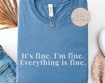 May include: A blue t-shirt with the text "It's fine. I'm fine. Everything is fine." A silver necklace with a sunburst pendant is on the shirt. The shirt is folded on a white surface.