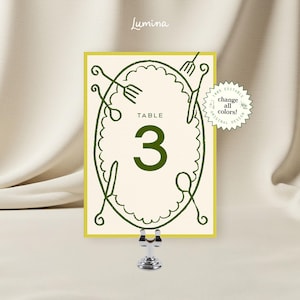 May include: A table number card with a cream background and a green border. The card features the number 3 in a large, bold font, and the word "TABLE" above it. Decorative cutlery surrounds the number. The card is held by a silver metal clip.