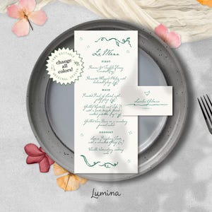 May include: A wedding menu and place card set on a gray plate, with a fork and knife. The menu features green script text, including "Le Menu" and a list of dishes. The place card has a name and a heart symbol. The design includes floral accents.