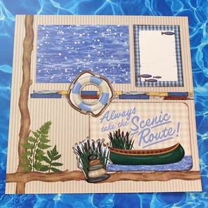 May include: A scrapbook page with a scenic lake theme. It features a blue and white water background, a life preserver, a green canoe, and the text "Always take the Scenic Route!" Decorative elements include fish, cattails, and a stack of stones.