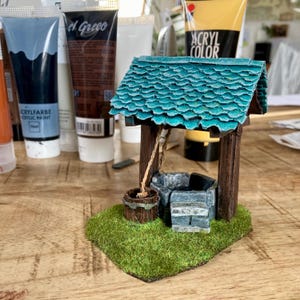 May include: A miniature wishing well model featuring a textured teal shingled roof, dark brown wooden support posts, and a stone-look base. A small brown bucket hangs from a rope, and the entire structure sits on a patch of green artificial grass.