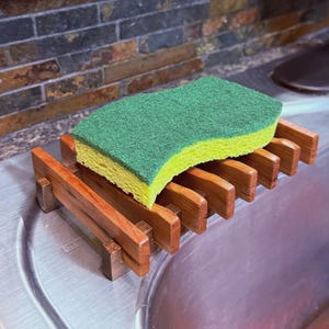 Puede incluir: Un soporte de madera para jabón de platos con una esponja verde y amarilla. El soporte es de madera marrón rojiza y tiene un diseño de listones. La esponja es rectangular y se asienta sobre el soporte, lista para usar.