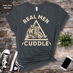 May include: Dark grey heather t-shirt with the text "REAL MEN CUDDLE" in a beige triangle, featuring a graphic of two figures in a martial arts embrace. Black and white trainers are visible on a wooden surface.